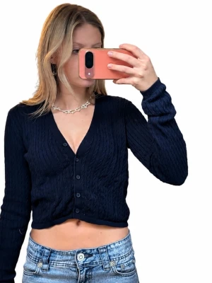 Mörkblå cropped kofta med knappar - Snygg mörkblå cropped kofta med kabelstickat mönster och v-ringning. Koftan har långa ärmar och stängs med fem knappar framtill. Perfekt att styla med jeans eller kjol för en trendig look.