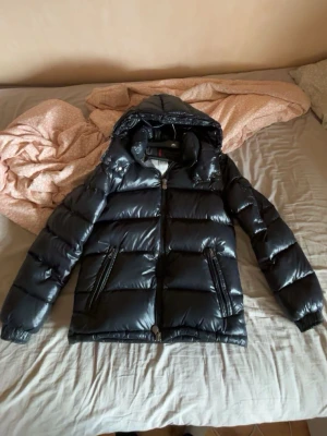 Svart dunjacka från Moncler (BUDA) - Snygg svart dunjacka från Moncler med glansig finish och klassisk Moncler-logga på bröstet. Jackan har huva, två dragkedjeförsedda fickor och quiltad design. Perfekt för kalla vinterdagar och riktigt trendig look.