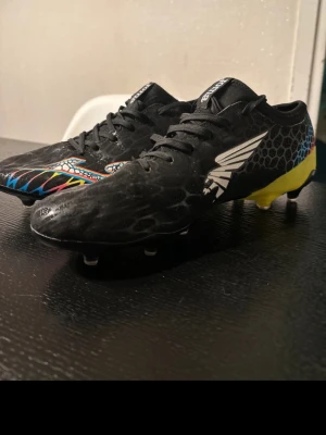 Svarta fotbollsskor med färgglada detaljer - Säljer ett par svarta fotbollsskor med coolt mönster och färgglada detaljer i blått, gult och rött. Skorna har snörning, låg profil och mönstrad ovandel i syntetmaterial. Perfekta för dig som vill sticka ut på planen.