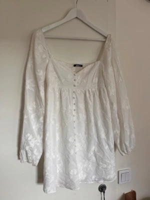 Vit blommig långärmad klänning  - Vit klänning från Gina Tricot, med långa ärmar och broderade blommor. Klänningen har knappar framtill med lätt rynk under bysten. Perfekt för sommaren.
