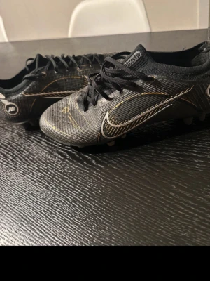 Svarta Nike fotbollsskor med guld - Säljer ett par svarta Nike fotbollsskor med snygga gulddetaljer och vita loggor. Skorna har snörning och dobbar för bra grepp på planen. Tillverkade i ett slitstarkt syntetmaterial som ger bra bollkontroll och passform.