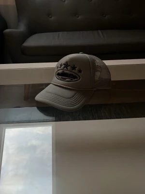 Beige truckerkeps med mesh och tryck - Säljer en beige truckerkeps med mesh på sidorna och bak, samt ett tryck med stjärnor och logga framtill. Kepsen har böjd skärm och justerbar snapback. Perfekt för en avslappnad streetwear-look. 