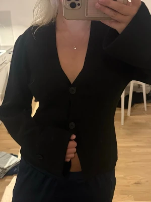 Svart kofta med knappar - Snygg svart kofta med två knappar framtill och djup V-ringning. Modellen är figurnära med långa ärmar. Storlek M från Gina tricot. Använd enstaka gång så är i mycket bra skick!
