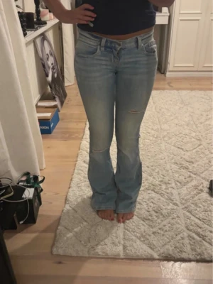 Ljusblå bootcut jeans med slitningar - Säljer ett par ljusblå bootcut jeans med lätt slitna detaljer på låret och knät. Jeansen har klassisk femficksmodell och normal midja. Perfekta för dig som gillar en avslappnad men trendig look.