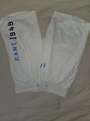 Vita shorts från Gant med snörning - Säljer ett par vita shorts från Gant med trycket 'GANT 1949' i svart och blått på ena benet. Shortsen har snörning i midjan och är gjorda i mjuk bomull, perfekta för chill dagar. Klassisk och avslappnad stil.