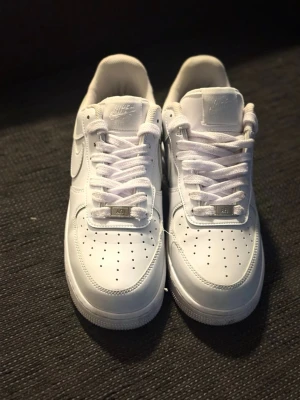 Nike Air Force 1 vita sneakers - Klassiska Nike Air Force 1 sneakers i helvitt läder med perforerad tå, vit sula och matchande vita skosnören. Ikonisk Swoosh-logga på sidan och metallbricka på snörningen. Perfekt för en clean och stilren look.
