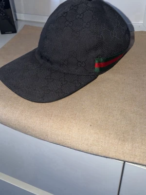 Svart gucci keps - Säljer en svart keps från Gucci med det klassiska GG-monogrammönstret över hela kepsen. På sidan finns en grön och röd rand som ger den en ikonisk touch. Kepsen är i tygmaterial och har böjd skärm. Spännet är lite avklippt där bak, dustbag följer med 