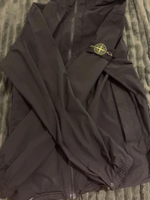 Svart vindjacka från Stone Island - Snygg svart vindjacka från Stone Island med klassisk logotyp på ärmen. Jackan har dragkedja framtill, två sidofickor och elastiska muddar vid ärmslut och nederkant. Perfekt för blåsiga dagar och enkel att matcha med olika outfits.