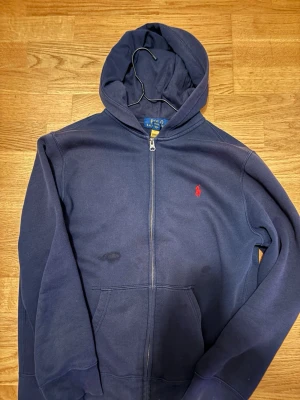 Mörkblå hoodie med dragkedja Polo - Mörkblå hoodie från Polo Ralph Lauren med dragkedja framtill och klassisk röd logga på bröstet. Tröjan har huva, kängurufickor och ribbade muddar vid ärmslut och nederkant. Perfekt för en avslappnad och stilren look.