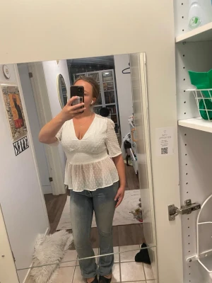 Vit prickig blus med v-ringning - Söt vit blus med v-ringning och korta ärmar. Blusen har ett lätt transparent material med små prickar och markerad midja som ger en fin siluett. Volang nedtill för extra detalj. Perfekt att styla med jeans eller kjol.