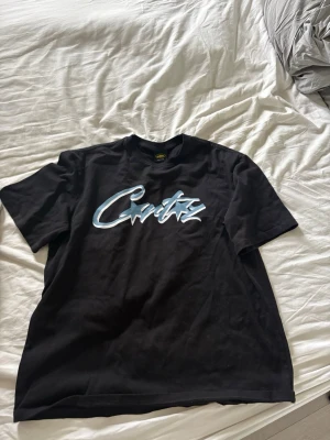 Corteiz T-shirt - Svart t-shirt med ett stort blåvitt grafiskt tryck på bröstet. Klassisk passform och rund halsringning, perfekt för en avslappnad streetwear-look.
