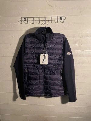 Mörkblå dunväst från Moncler - Snygg mörkblå dunväst från Moncler med quiltad front och stickade ärmar. Västen har dragkedja framtill, två fickor med dragkedja och Moncler-logga på ärmen. Perfekt för lager-på-lager och riktigt trendig look.