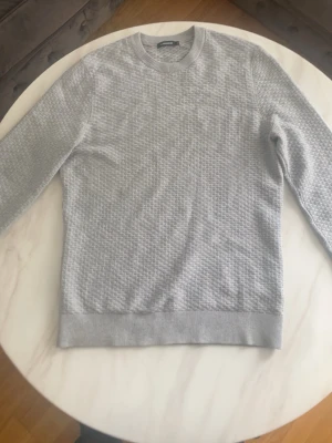 Ljusgrå tröja från J.Lindeberg - Snygg ljusgrå tröja från J.Lindeberg med diskret rutmönster. Tröjan har rund halsringning, långa ärmar och ribbade muddar vid ärmslut och nederkant. Perfekt för dig som gillar stilrena och klassiska plagg.