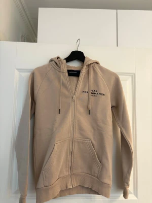 Hoodie Peak Performance - Beige hoodie från Peak Performance med dragkedja, huva och fickor framtill. Mjuk fleece på insidan och diskret logga på bröstet.