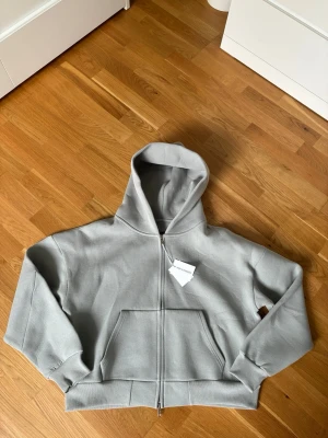 Saint Alexander zip-up hoodie  - Snygg ljusgrå hoodie från Saint Alexander med dragkedja framtill och stor känguruficka. Tillverkad i mjukt bomullsmaterial och har en klassisk huva. Perfekt för en avslappnad och trendig look.