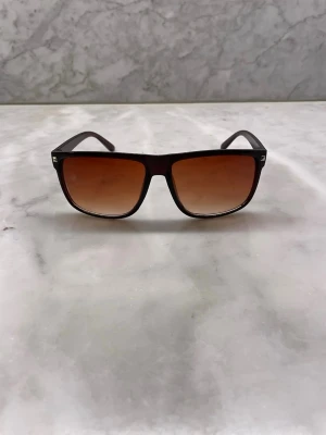 Solglasögon  - Snygga solglasögon med bruna bågar och fyrkantig form. Glasen har en brun toning som ger en cool retrovibe. Passar perfekt till dig som vill ha en stilren accessoar med lite attityd. Tillverkade i plast.