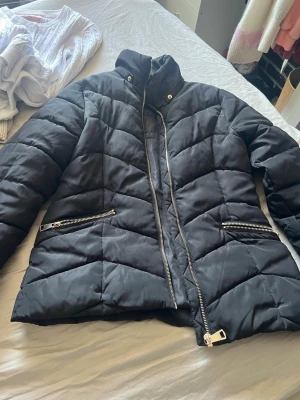 Svart pufferjacka från KappAhl - Snygg svart pufferjacka från KappAhl med hög krage och dragkedja framtill. Jackan har två fickor med silverfärgade dragkedjor och ett quiltat mönster som ger en cool look. Perfekt för kalla vinterdagar och riktigt skön att bära.