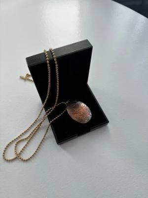 Silverfärgat ovalt medaljonghalsband - Säljer ett silverfärgat halsband med en oval medaljong som har graverade blommönster. Kedjan består av små pärlor och medaljongen öppnas för att lägga in ett foto eller minne. Perfekt accessoar för dig som gillar vintage och unika detaljer.