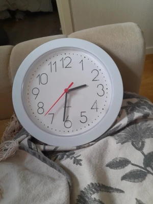 Vit rund väggklocka - Säljer en stilren rund väggklocka i vit plast med svart urtavla och siffror. Klockan har svarta visare för timmar och minuter samt en röd sekundvisare. Enkel och modern design som passar i de flesta rum.