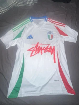 Italien special Stüssy x Adidas - Säljer en vit Italien matchtröja i samarbete mellan Stüssy och Adidas. Tröjan har gröna och röda detaljer på sidorna och axlarna, blå Adidas-logga och Italiens landslagsemblem på bröstet. Stüssy-loggan i rött framtill. Tillverkad i lätt och ventilerande material.