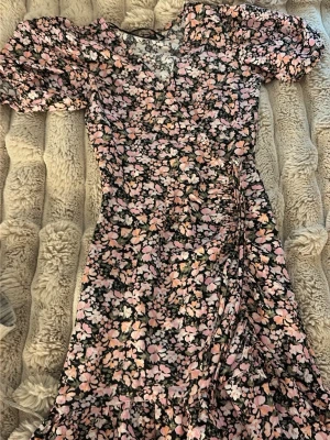 Blommig omlottklänning med volang - Söt blommig omlottklänning i svart, rosa och beige med volangkant och korta puffärmar. Klänningen har v-ringning och knyt i midjan för en snygg passform. Perfekt för sommarens alla tillfällen.