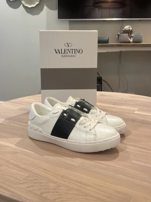 Valentino Garavani vita sneakers - Snygga vita sneakers från Valentino Garavani med bred svart läderdetalj över vristen och nitar på sidan. Skorna har en klassisk låg profil. Perfekta för dig som vill ha en stilren och exklusiv look.