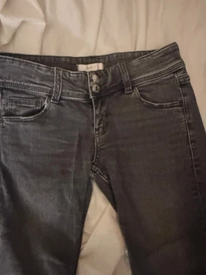 Vintage svarta jeans från Gina Tricot - Ett par svarta jeans från Gina Tricot med två synliga knappar i midjan i bootcut och klassisk femficksmodell. Jeansen har en smal passform och är tillverkade i stretchigt denimtyg för extra komfort. Perfekta för en trendig och avslappnad look.
