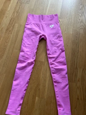 Rosa sömlösa leggings - Snygga rosa leggings med hög midja och sömlös design. Materialet är stretchigt och har diskret struktur längs benen. På höften finns en vit logga med en siluett av ett ansikte. Perfekta för träning eller chill.