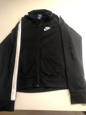 Svart zip-hoodie från Nike - Snygg svart zip-hoodie från Nike med vit logga på bröstet och en vit rand längs ena ärmen. Tröjan har huva, dragkedja framtill och två fickor. Perfekt till träning eller chill. Tillverkad i mjukt material för extra komfort.