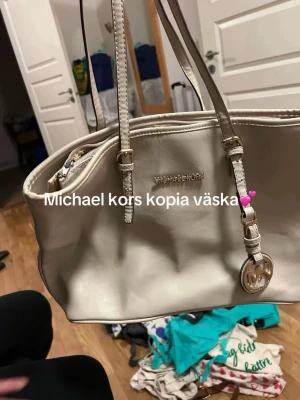Beige handväska med gulddetaljer - Stilren beige handväska i skinnimitation med två långa axelband och guldfärgade detaljer. Väskan har ett hängande MK-emblem och dragkedja upptill. Perfekt för dig som gillar klassisk design och vill ha plats för det viktigaste.