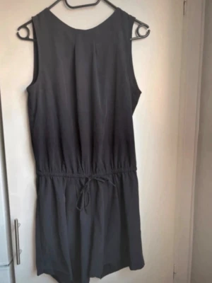 Svart ärmlös jumpsuit med knyt - Säljer en svart ärmlös jumpsuit med knyt i midjan och öppen ryggdetalj. Supersnygg, passar bara inte mig. Aldrig använd!