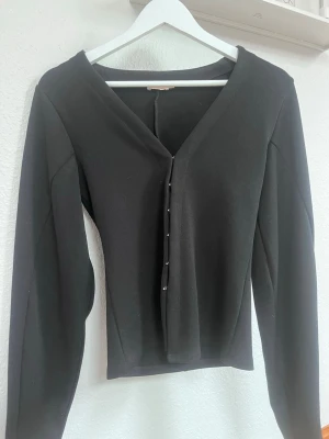 Svart v-ringad topp med knappar - Stilren svart topp med v-ringning och knäppning framtill från Nelly. Långärmad modell med figurnära passform som ger en snygg siluett. Perfekt att matcha med både jeans och kjol för en trendig look.❣️