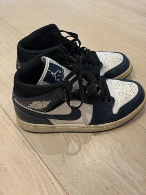 Air Jordan 1 Mid blå/vit - Snygga Air Jordan 1 Mid sneakers i blått och vitt med svarta detaljer. Skorna har klassisk Jordan-logga på sidan och tungan, samt svart snörning. Ovandelen är i läder och sulan är vit med beige kant. Perfekta för dig som vill ha en ikonisk streetwear-look.