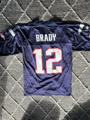Mörkblå Patriots NFL-tröja nr 12 Tom Brady - Säljer en mörkblå New England Patriots NFL-tröja med nummer 12 och namnet BRADY på ryggen. Tröjan har vita och röda detaljer, korta ärmar och meshmaterial för extra ventilation. Perfekt för dig som älskar amerikansk fotboll och vill representera ditt lag eller som samlar objekt. Bra skick.