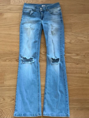 Ljusblå bootcut jeans med slitningar - Säljer ett par ljusblå bootcut jeans med slitna detaljer över knäna och klassiska fem fickor. Jeansen har en låg midja och utställda ben, samt snygga bakfickor med lock och knapp. Perfekta för en avslappnad och trendig look.