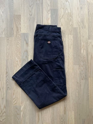 Dickies Work Pants - W32 L30, mörkblå - OBS - Upplevd storlek, mått i beskrivningen  Bra skick. Få tecken på användning Byxor från Dickies, upplevd storlek W32 L30 (mått nedan), mörkblå Logga höger bakficka, detaljfickor bak/sida höger, knapp med dragkedjegylf  Ej elastisk midja, uppskattad storlek W32 874-stil.  Midjemått - 84 cm Beninnerlängd - 77 cm (≈L30) Benvidd lår - 57 cm Benvidd knä - 47 cm Benvidd benslut - 46 cm  Byxorna är uppsydda och måtten är anpassade efter det.   Etikett bortklippt. Bild bevisar detta  Inga defekter