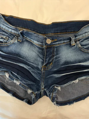 Blå lågmidjade slitna jeansshorts - Säljer ett par blå jeansshorts med slitna detaljer och råa kanter. Shortsen har klassisk femficksdesign, knappgylf och kontrastsömmar. Perfekta för sommaren och ger en avslappnad vibe.
