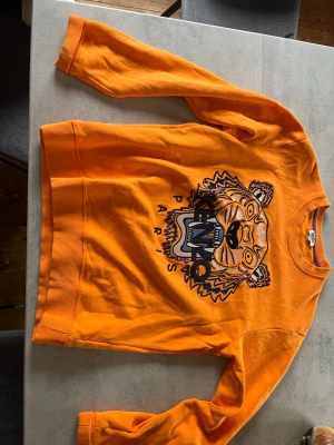 Kenzo tröja  - orange sweatshirt från Kenzo med det ikoniska tigerhuvudet broderat framtill och texten 'Kenzo Paris'. Tröjan har rund hals, ribbade muddar och är tillverkad i mjuk bomull. Perfekt för dig som vill sticka ut med en färgstark look.