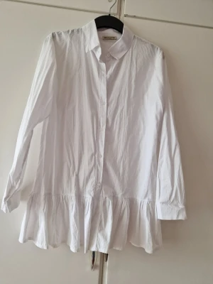 Vit skjorta med volangkant - Säljer en vit skjorta med klassisk krage, knäppning framtill och lång ärm. Skjortan har en snygg volangkant nedtill som ger en trendig och lekfull look. Perfekt att styla med jeans eller kjol för en cool outfit.