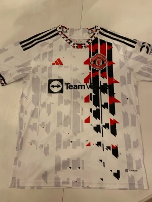 Manchester United bortatröja M - Säljer en Manchester United bortatröja från Adidas i storlek M. Tröjan är vit med svart och rött grafiskt mönster, klubbmärke på bröstet och TeamViewer-logga. Slim fit-modell med korta ärmar och klassiska Adidas-ränder på axlarna. Perfekt för fotbollsträning eller match.