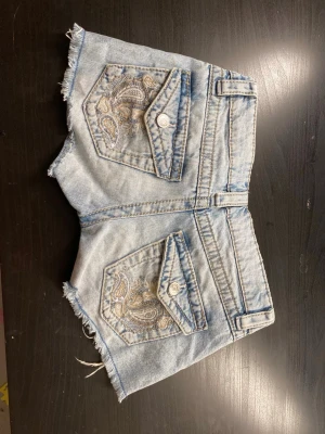 Ljusblå jeansshorts med broderade fickor - Säljer ett par ljusblå jeansshorts med råa kanter och snygga broderade bakfickor. Shortsen har normal passform, bälteshällor och knappstängning framtill. Perfekta för sommardagar och ger en avslappnad vibe.