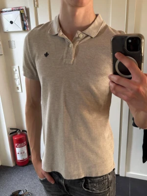 Morris Beige Piké - Snygg beige pikétröja med klassisk krage och knappar framtill. Tröjan har korta ärmar och en liten svart broderad detalj på bröstet. Perfekt till jeans eller shorts för en clean och enkel stil. Hör av dig vid frågor!