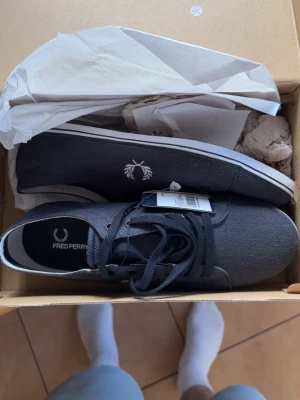 Mörkblå Fred Perry canvas sneakers - Snygga mörkblå sneakers från Fred Perry i canvasmaterial med vita detaljer och klassisk logga på sidan. Skorna har låga skaft, svarta snören och vit sula för en clean och stilren look. Köpta för 1899kr säljs för 1,100kr pga min farbror köpte fel storlek till mig i födelsedagspresent.