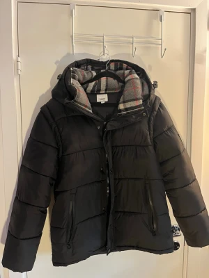 Svart pufferjacka från Burberry - Snygg svart pufferjacka från Burberry med huva och dragkedja. Jackan har två fickor med dragkedja och ett klassiskt rutigt foder i grått, vitt och rött. Perfekt för kalla dagar och riktigt varm tack vare det tjocka materialet.