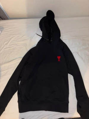 Svart hoodie från Ami Paris - Svart hoodie från Ami Paris med klassisk huva och känguruficka. På bröstet finns Ami:s ikoniska röda hjärta med A-bokstav. Tillverkad i mjukt bomullsmaterial, perfekt för en avslappnad och stilren look.