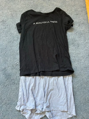 Svart pyjamas-set med text - Mysigt pyjamas-set med svart t-shirt och vit text 'A BEAUTIFUL MESS' på bröstet samt ljusgrå shorts. T-shirten har korta ärmar och rund hals. Materialet ser ut att vara mjukt och bekvämt, perfekt för slappa dagar eller nätter.