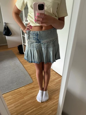 Kort jeanskjol - Säljer en ljusblå kort jeanskjol med plisserad nederdel och ett coolt bältdetalj i midjan. Kjolen har en avslappnad passform och är perfekt för dig som gillar en trendig och avslappnad stil.