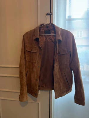 Brun skinnjacka från Jofama - Snygg brun skinnjacka från Jofama i mocka-look. Jackan har två bröstfickor med lock och knappar, samt två snedställda fickor framtill. Klassisk krage och knappstängning framtill ger en tidlös stil.