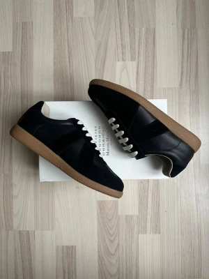 Svarta sneakers från Maison Margiela - Stilrena svarta sneakers från Maison Margiela med vita skosnören och klassisk brun gummisula. Skorna har en låg profil och är tillverkade i en mix av läder och mocka för en snygg kontrast. Perfekta för dig som gillar minimalistisk och tidlös design.