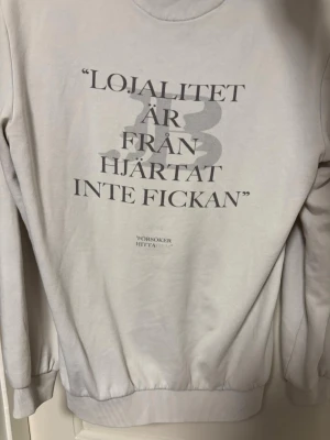 JAFFAR BYNS EGNA MERCH - Vit hoodie med tryck på bröstet och texten 'Lojalitet är från hjärtat inte fickan' på ryggen. Har en klassisk känguruficka och huva. Materialet ser ut att vara mjukt bomullstyg, perfekt för en avslappnad stil. Tröjan är efterfrågad och säljs inte längre! Står ej storlek men skulle uppskatta den som S!
